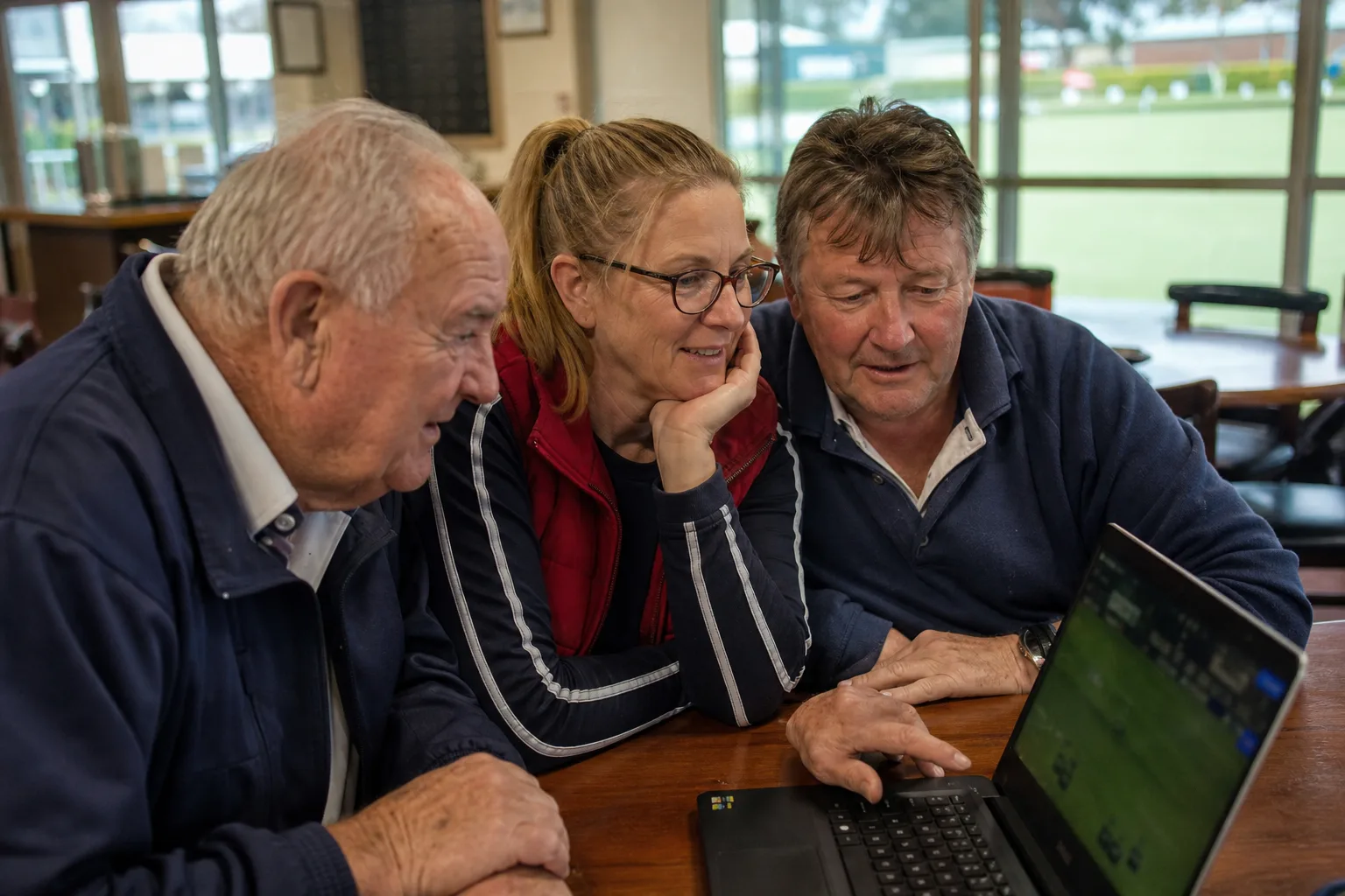 Three bowls club members gathered around a laptop playing a digital lawn bowling game